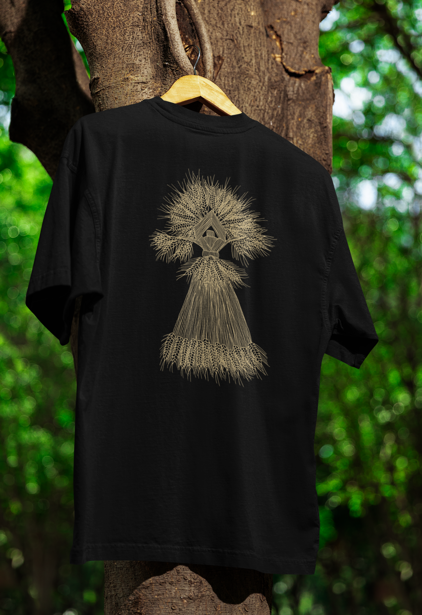 A black T-shirt with an golden coloured illustration of a corn dolly on the front hanging on a tree.